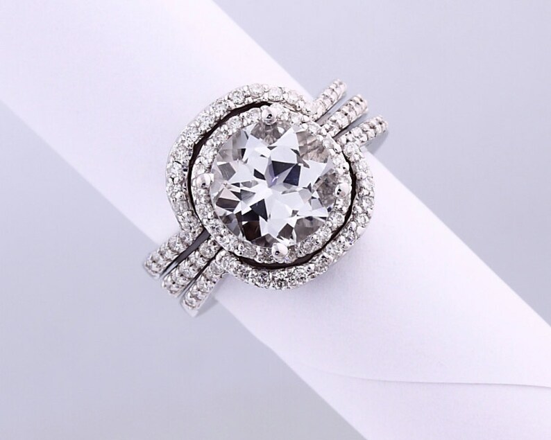 May include: A white gold engagement ring with a large, round, clear center stone surrounded by a halo of smaller diamonds. The ring is set with a double band of smaller diamonds.
