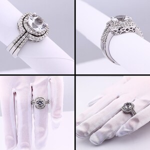 May include: A silver engagement ring set with a large, clear, oval-shaped center stone surrounded by smaller stones. The ring is on a white background.