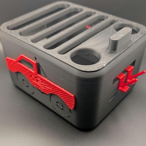 May include: A black and red 3D-printed desk organizer. The organizer features a truck design on the side and slots on top for pens and other supplies. It has a small red arrow design on the side.