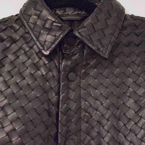 May include: Close-up of a black woven leather jacket. The jacket features a collared neckline and button-down front. The intricate weave pattern creates a textured surface, and the jacket appears to be made of high-quality leather.