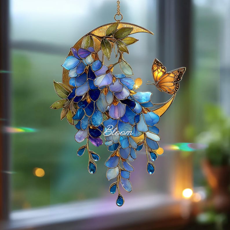 Peut inclure: Un attrape-soleil en vitrail repr&eacute;sentant un croissant de lune dor&eacute; orn&eacute; de fleurs de glycine bleues en cascade, de feuilles vertes et d'un papillon orange. Le mot Bloom est &eacute;crit en lettres blanches au centre de la composition florale.