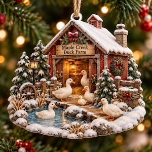 Peut inclure: Une décoration de Noël suspendue en forme de grange rouge au toit enneigé, portant l'inscription Maple Creek Duck Farm. La scène montre cinq canards blancs, un petit étang, des sapins, une lanterne et une couronne, le tout saupoudré de neige.