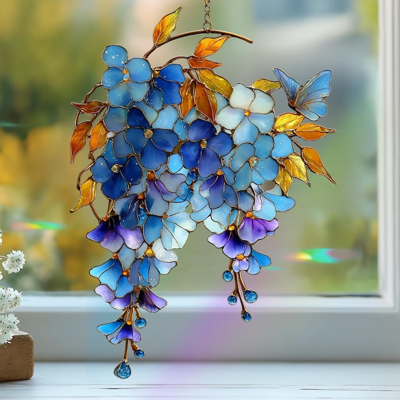 Peut inclure: Un attrape-soleil en vitrail repr&eacute;sentant un bouquet de fleurs bleues et violettes avec des feuilles ambr&eacute;es. Le motif comprend un petit papillon bleu pos&eacute; sur le c&ocirc;t&eacute; droit, avec une fine armature m&eacute;tallique soulignant chaque p&eacute;tale.