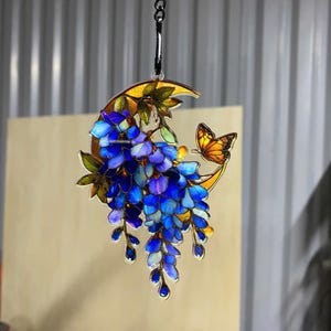 Peut inclure: Une d&eacute;coration suspendue en forme de croissant de lune aux tons ambr&eacute;s, orn&eacute;e de fleurs de glycine bleues en cascade et d'un petit papillon orange. L'objet est r&eacute;alis&eacute; dans un style vitrail et suspendu par une cha&icirc;ne en m&eacute;tal argent&eacute;.