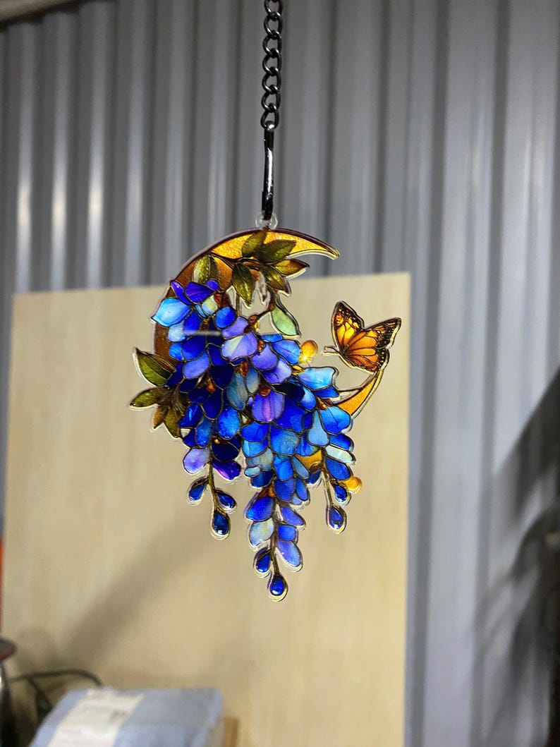 Peut inclure: Une d&eacute;coration suspendue en forme de croissant de lune aux tons ambr&eacute;s, orn&eacute;e de fleurs de glycine bleues en cascade et d'un petit papillon orange. L'objet est r&eacute;alis&eacute; dans un style vitrail et suspendu par une cha&icirc;ne en m&eacute;tal argent&eacute;.