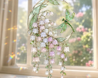 Peut inclure: Un attrape-soleil suspendu composé d'un cadre en forme de croissant de lune avec des feuilles vertes translucides, des fleurs de muguet rose pâle et blanc, et une libellule verte aux ailes délicates, le tout avec des contours métalliques.