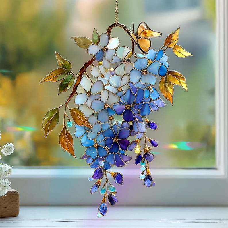 Peut inclure: Un attrape-soleil en vitrail repr&eacute;sentant une grappe de fleurs bleues, violettes et blanches sur une branche avec des feuilles ambr&eacute;es. Un petit papillon repose sur la branche sup&eacute;rieure. L'objet est suspendu par une fine cha&icirc;ne m&eacute;tallique.