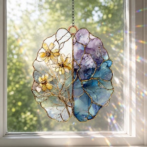 May include: A brain-shaped stained glass suncatcher hanging by a metal chain. The left side features yellow flowers on thin branches against a clear background, while the right side displays marbled shades of purple and blue glass.