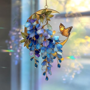 Peut inclure: Un attrape-soleil en vitrail repr&eacute;sentant un croissant de lune orn&eacute; de fleurs de glycine bleues et violettes en cascade, de feuilles vertes et d'un papillon orange aux motifs d'ailes noirs, suspendu par une fine cha&icirc;ne m&eacute;tallique.