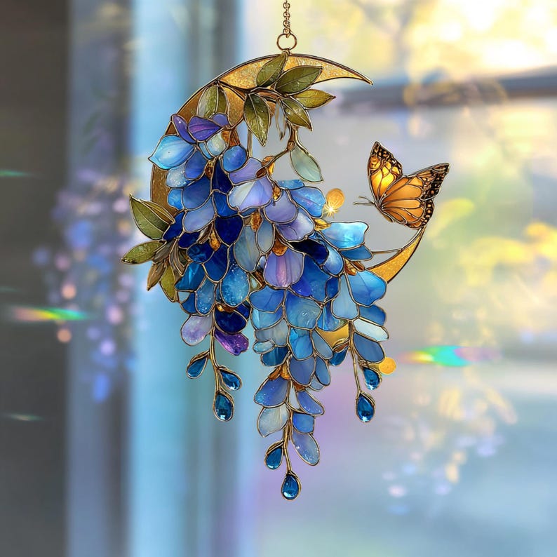 Peut inclure: Un attrape-soleil en vitrail repr&eacute;sentant un croissant de lune orn&eacute; de fleurs de glycine bleues et violettes en cascade, de feuilles vertes et d'un papillon orange aux motifs d'ailes noirs, suspendu par une fine cha&icirc;ne m&eacute;tallique.