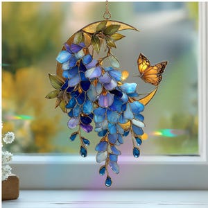 Peut inclure: Un attrape-soleil en vitrail repr&eacute;sentant un croissant de lune dor&eacute; orn&eacute; de fleurs de glycine bleues et violettes en cascade, de feuilles vertes et d'un papillon orange. L'objet est suspendu par une fine cha&icirc;ne dor&eacute;e aux contours m&eacute;talliques.