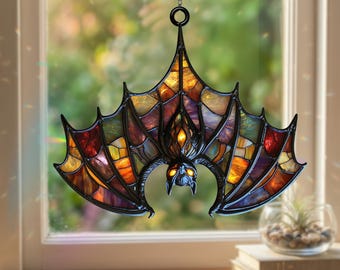Könnte beinhalten: Ein Fledermaus-Sonnenfänger aus Buntglas mit einem schwarzen Metallrahmen und mehrfarbigen Glasscheiben in Bernstein-, Orange-, Violett- und Rottönen. Die Fledermaus hängt an einer Metallkette und hat weit ausgebreitete Flügel.