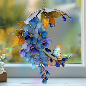 Peut inclure: Un attrape-soleil en vitrail en forme de croissant de lune orn&eacute; de fleurs bleues et violettes, de feuilles jaune dor&eacute; et d'un petit papillon. L'objet est assembl&eacute; avec une fine structure m&eacute;tallique et des accents en verre bleu.