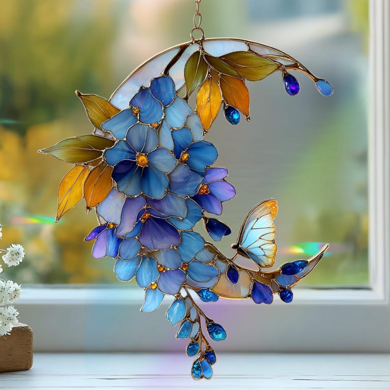 Peut inclure: Un attrape-soleil en vitrail en forme de croissant de lune orn&eacute; de fleurs bleues et violettes, de feuilles jaune dor&eacute; et d'un petit papillon. L'objet est assembl&eacute; avec une fine structure m&eacute;tallique et des accents en verre bleu.