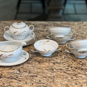 May include: A white porcelain tea set with gold trim and wheat-like designs. The set includes a sugar bowl with a lid, a gravy boat, soup bowls with handles, and saucers. The pieces are arranged on a speckled countertop.