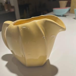 May include: A pale yellow ceramic creamer with a handle and a spout. The creamer has a ribbed design and a glossy finish. The interior is a slightly darker shade of yellow. The creamer is sitting on a white surface.