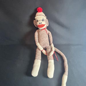 May include: A sock monkey doll crafted from brown and cream-colored knit fabric. The doll features a red pom-pom on its head, black button eyes, stitched eyelashes, and a bright red mouth. A thin red ribbon is tied around its neck and wrists.