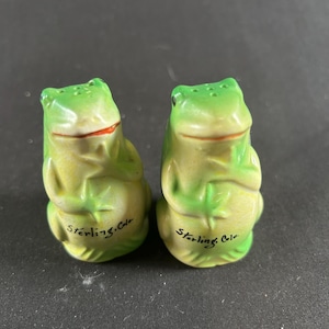 May include: A pair of ceramic frog-shaped salt and pepper shakers. The frogs are primarily green with yellow bellies and red mouths. The text "Sterling, Colo" is printed on the base of each shaker.