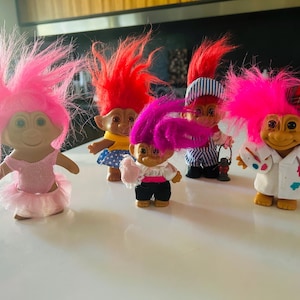 May include: A collection of vintage Troll dolls with vibrant pink, red, and purple hair. The dolls are dressed in various outfits, including a tutu, sailor suit, and artist's smock. They are posed on a white surface.