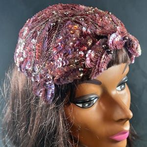 May include: A decorative cap covered in shimmering mauve and purple sequins, accented with intricate silver beadwork and small dark spherical beads. The headpiece features a textured, layered design that covers the crown of the head.