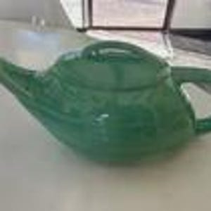 May include: A glossy, jade green ceramic teapot with a curved spout and a matching lid. The teapot has a rounded body and a loop handle. The teapot is sitting on a white surface.