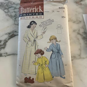 May include: A Butterick printed sewing pattern envelope 6234 for a robe, featuring illustrations of a person in a white and yellow plaid robe, a child in a yellow robe, and a child in a blue robe. The text includes Breast 24, Size 6, and 35 cents.