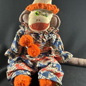 May include: Handmade sock monkey doll wearing a clown outfit. The monkey has a beige sock body, orange mouth, and green button eyes. It wears a blue, red, and beige patterned outfit with orange pom-poms and feet. The hat is beige with an orange brim.