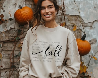 May include: A beige sweatshirt with the word "fall" in a cursive font. The sweatshirt has a list of fall-themed words, including "cozy sweaters," "pumpkin patch," and "hayrides." The background includes pumpkins and a stone wall.