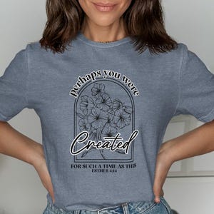May include: A blue-grey t-shirt with the words "perhaps you were created" above a floral design. Below the design, the text reads "for such a time as this, Esther 4:14".
