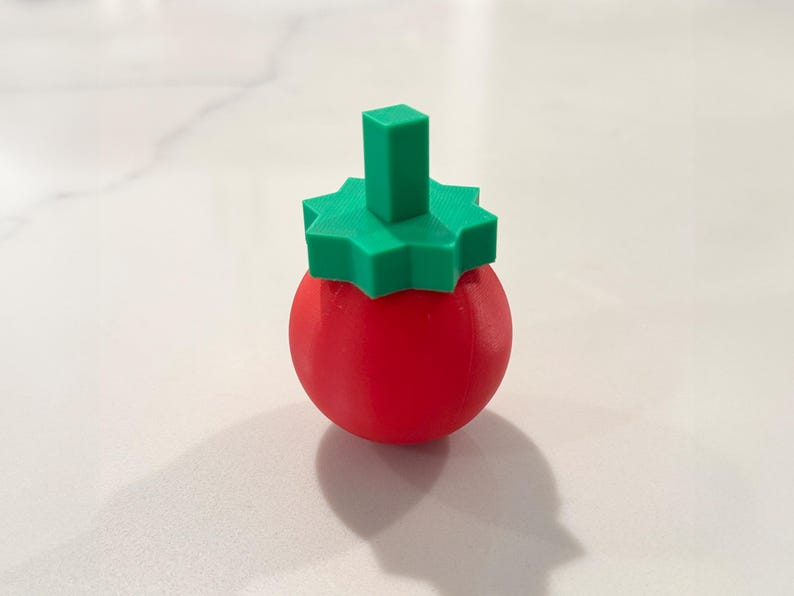 Op de afbeelding: Een rood, bolvormig object met een groene, stervormige bovenkant en een rechthoekige steel. Het object is waarschijnlijk een decoratief item of een unieke flesstopper, tegen een witte achtergrond. 3D-geprint.