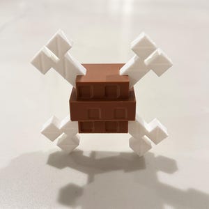 Op de afbeelding: Een 3D-geprint speelgoed dat een doodshoofd met gekruiste botten voorstelt. De centrale structuur is bruin, met een rechthoekige vorm. De botten zijn wit en steken naar buiten uit. Het speelgoed heeft een pixelachtige esthetiek.