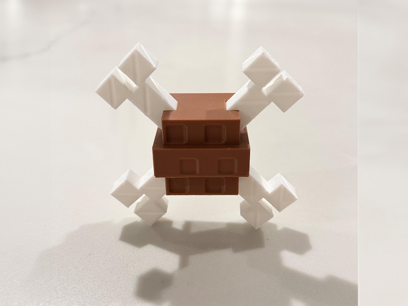 Op de afbeelding: Een 3D-geprint speelgoed dat een doodshoofd met gekruiste botten voorstelt. De centrale structuur is bruin, met een rechthoekige vorm. De botten zijn wit en steken naar buiten uit. Het speelgoed heeft een pixelachtige esthetiek.
