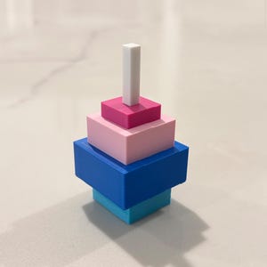 Op de afbeelding: Een kleurrijk, geometrisch stapelspeelgoed. De basis is een lichtblauwe vierkant, bekroond door een donkerblauwe vierkant, een roze vierkant, een magenta vierkant en een wit rechthoekig prisma. Het speelgoed is ontworpen om te stapelen.