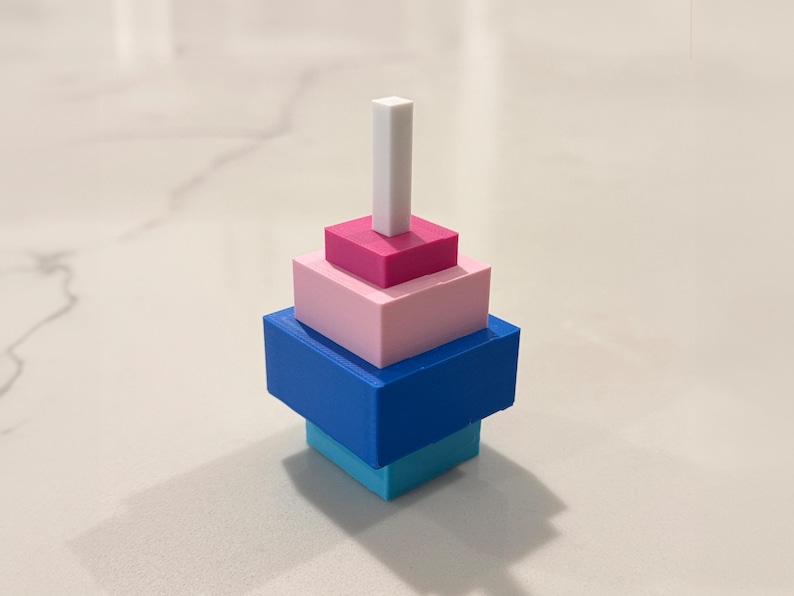 Op de afbeelding: Een kleurrijk, geometrisch stapelspeelgoed. De basis is een lichtblauwe vierkant, bekroond door een donkerblauwe vierkant, een roze vierkant, een magenta vierkant en een wit rechthoekig prisma. Het speelgoed is ontworpen om te stapelen.