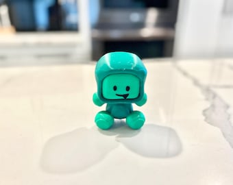 May include: A small, teal-colored figurine of a smiling character wearing a helmet. The figure has a square face, round body, and ball-shaped feet and hands. The figurine is sitting on a white surface.