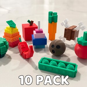Op de afbeelding: Een verzameling van tien kleurrijke, geometrische voedselminiaturen. Inclusief een groene appel, een rode peper, een wortel, een tomaat, een kokosnoot en een bakje groene erwten. De tekst "10 PACK" staat onderaan.