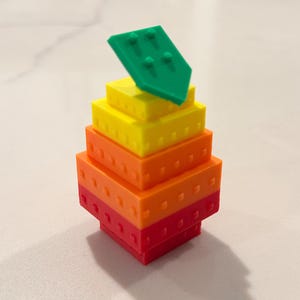 Op de afbeelding: Een kleurrijk, geometrisch speelgoed gemaakt van gestapelde blokken. De basis is rood, gaat over in oranje, geel en wordt bekroond met een groen stuk. De blokken hebben kleine, cirkelvormige uitsteeksels. Dit speelgoed is ontworpen om te bouwen en creatief te spelen.
