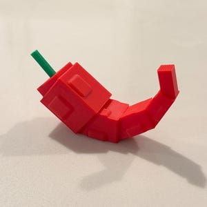 Op de afbeelding: Een 3D-geprinte rode chilipeper met een groene steel. De peper is gemaakt van geometrische vormen en heeft een gebogen ontwerp. De peper ligt op een wit oppervlak.