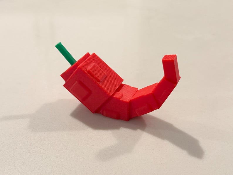 Op de afbeelding: Een 3D-geprinte rode chilipeper met een groene steel. De peper is gemaakt van geometrische vormen en heeft een gebogen ontwerp. De peper ligt op een wit oppervlak.