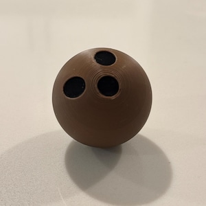 Op de afbeelding: Een bruine bowlingbal met drie zwarte vingergaten. De bal is bolvormig en ligt op een lichtgekleurd oppervlak, waardoor een schaduw ontstaat. De bowlingbal is een klassiek sportartikel.