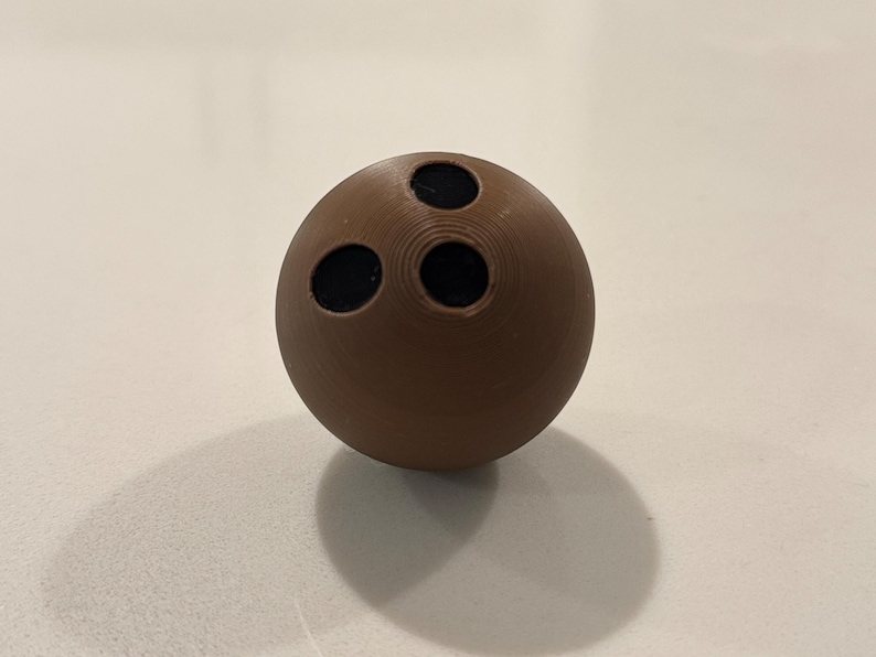 Op de afbeelding: Een bruine bowlingbal met drie zwarte vingergaten. De bal is bolvormig en ligt op een lichtgekleurd oppervlak, waardoor een schaduw ontstaat. De bowlingbal is een klassiek sportartikel.