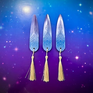 May include: Three blue and white decorative bookmarks with a floral design and a gold tassel. The bookmarks are arranged vertically against a starry blue and purple background.