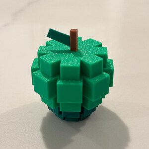 Op de afbeelding: Een groen, gepixeld appelvormig object met een bruine steel en een groen blad. Het object is gemaakt van gestapelde, vierkante blokken. De appel staat op een wit oppervlak.