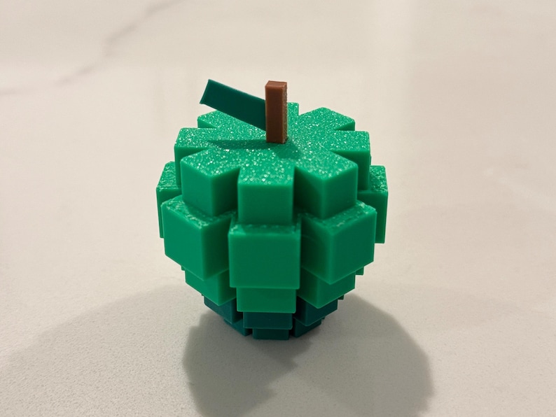 Op de afbeelding: Een groen, gepixeld appelvormig object met een bruine steel en een groen blad. Het object is gemaakt van gestapelde, vierkante blokken. De appel staat op een wit oppervlak.