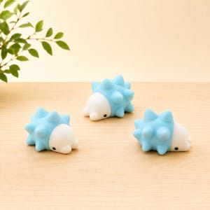 May include: Three small, spiky figurines shaped like hedgehogs. Each figurine features a smooth white face with a tiny black eye and a light blue body covered in rounded, soft-looking spikes. The three figures are arranged on a flat surface.