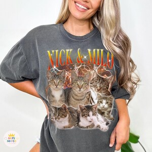 May include: Dark gray t-shirt featuring a graphic of seven cats with the text "NICK & MILO" in a retro font. The design includes lightning bolts and a vintage aesthetic, perfect for cat lovers.
