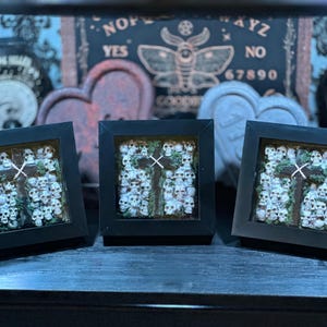 May include: Three framed artworks with black frames. Each piece displays a cross design surrounded by white skulls. The background includes a Ouija board and other gothic decor, creating a dark aesthetic.
