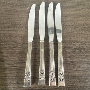 Pode incluir: Quatro facas de manteiga prateadas com cabos ornamentados. Os cabos têm um design canelado com um padrão floral decorativo na base. As lâminas são lisas e ligeiramente curvas.