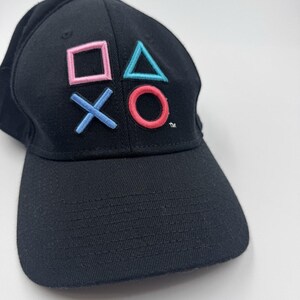 Puede incluir: Gorra de béisbol negra con el logotipo de PlayStation bordado en rosa, azul y turquesa. La gorra tiene una visera curva y un botón en la parte superior. El logotipo incluye un cuadrado, un triángulo, un círculo y una X.