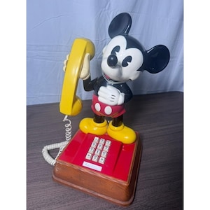 May include: A vintage Mickey Mouse telephone. The phone features a red base with a wooden-look trim, a yellow handset, and a Mickey Mouse figure. The phone has a white cord and a keypad with numbers and symbols.
